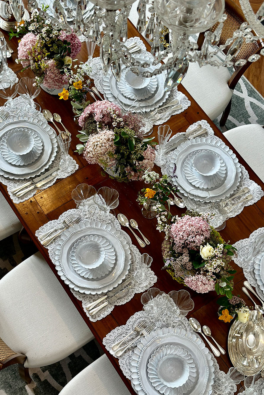 Elegant table setting with white Peggy plates, silverware, lace placemats and floral arrangements under a chandelier.