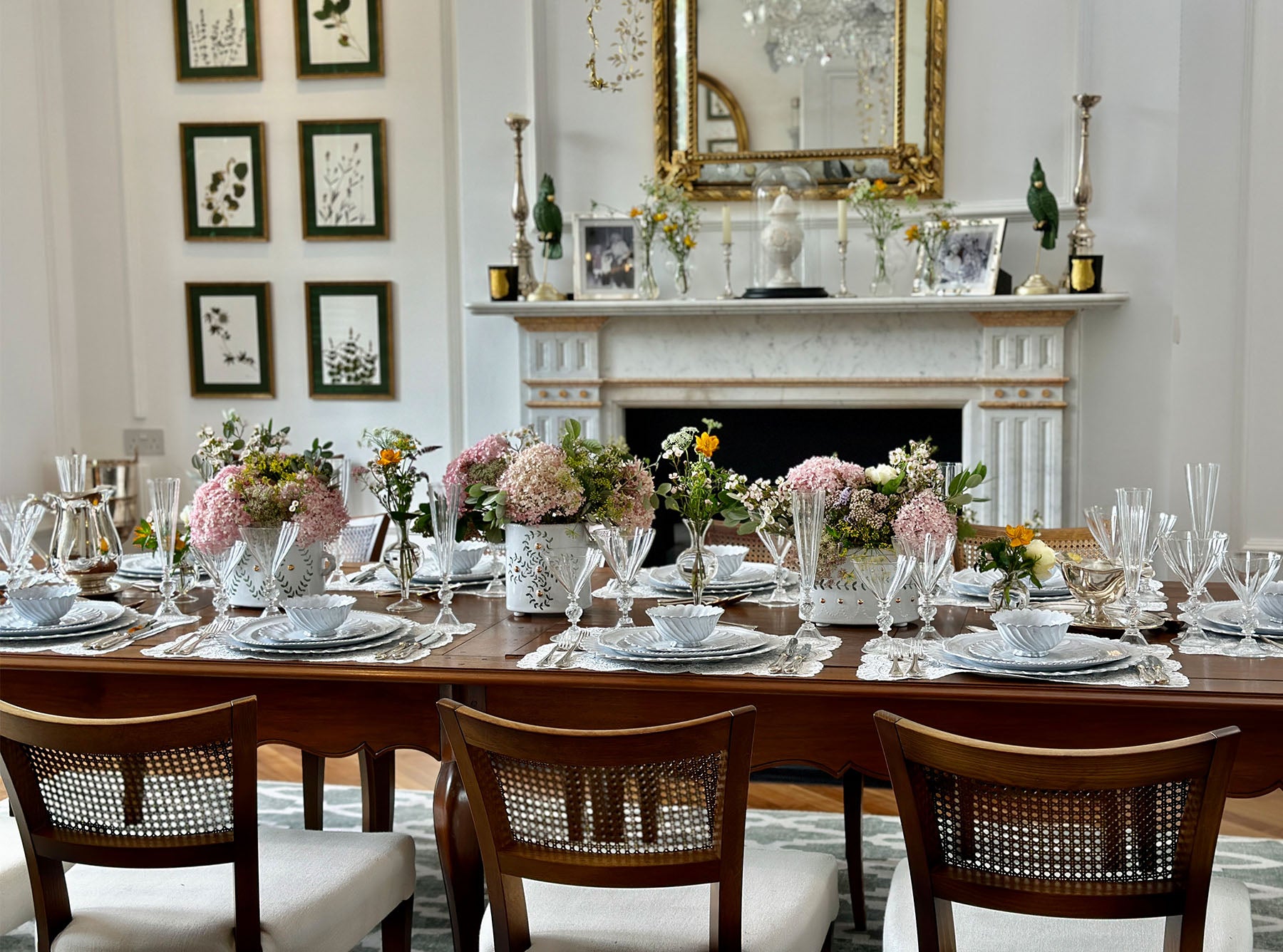 Dining room with a long wooden table set for a meal, featuring flowers, lace placemats and elegant ceramics.